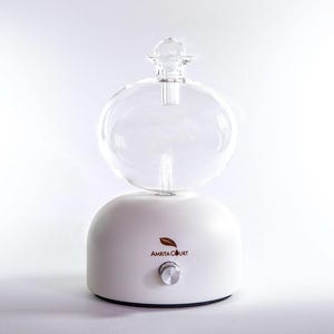 Op de afbeelding: Witte etherische olie diffuser met een heldere glazen bol en een kleine, ronde glazen bovenkant. De basis heeft een zilveren knop en de woorden "AMRITA COURT" met een bladlogo.