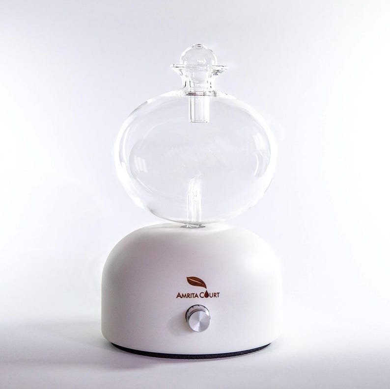 Op de afbeelding: Witte etherische olie diffuser met een heldere glazen bol en een kleine, ronde glazen bovenkant. De basis heeft een zilveren knop en de woorden "AMRITA COURT" met een bladlogo.