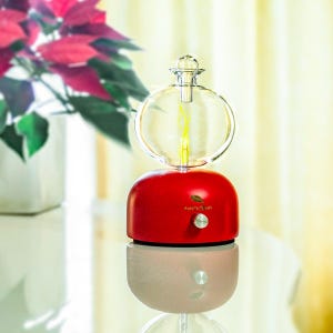 Op de afbeelding: Een rode en heldere glazen aromatherapie-diffuser met een ronde glazen bovenkant en een rode basis. De diffuser heeft een zilveren knop en de tekst "AMATA QURI" is op de voorkant gedrukt. De diffuser staat op een reflecterend oppervlak.