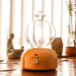 Op de afbeelding: Een heldere glazen etherische olie diffuser met een bolvormige bovenkant en een houten basis. De basis heeft een zilveren knop en de tekst "AMRITA COURT" met een bladlogo. De diffuser staat op een houten tafel.