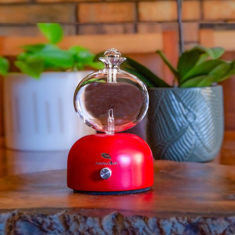 Op de afbeelding: Een rode aromatherapie diffuser met een heldere glazen bol en een kleine knop. De diffuser heeft de tekst "AMRITA LUXURY" en een bladlogo. De diffuser staat op een houten oppervlak, met planten op de achtergrond.