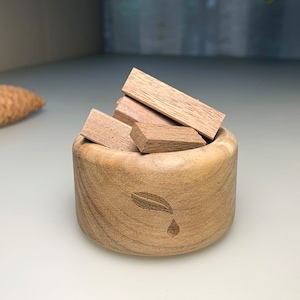 Puede incluir: Un cuenco de madera con un diseño de hoja y una gota de agua, lleno de pequeños palitos de madera para aromaterapia.