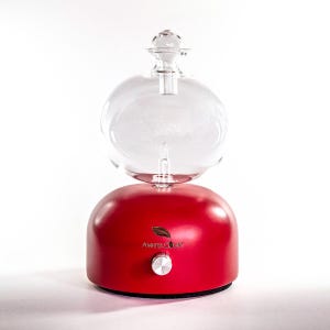 Op de afbeelding: Een rode etherische olie diffuser met een heldere glazen bol erbovenop. De basis heeft een zilveren knop en de tekst "AMRITA COURT". De diffuser is ontworpen om etherische oli&euml;n te verspreiden.
