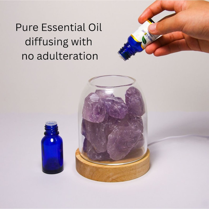 K&ouml;nnte beinhalten: Ein Glasdiffusor mit Holzsockel und einer klaren Glaskuppel, gef&uuml;llt mit Amethystkristallen. Eine blaue Glasflasche mit &auml;therischem &Ouml;l wird in den Diffusor gegossen. Der Text "Pure Essential Oil diffusing with no adulteration" ist im Bild sichtbar.