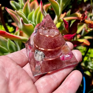 May include: A clear, pyramid-shaped orgone device with a reddish-pink stone center, copper wire coil, and small, colorful stones. The pyramid is held in a hand, with a succulent plant in the background.