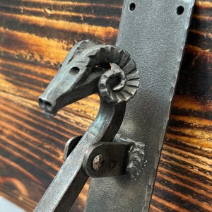 Pode incluir: Puxador de porta em metal forjado à mão com design de cabeça de carneiro. O puxador é fixado a uma placa retangular com orifícios de montagem. O metal tem um acabamento escuro e texturizado. O fundo é uma superfície de madeira com grão visível.