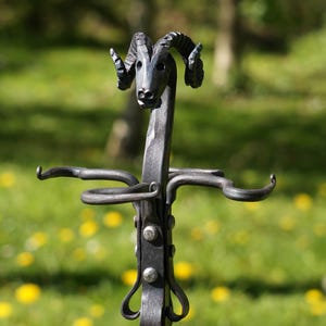 May include: A wrought iron garden torch stand with a ram's head design. The dark grey metal stand has curved arms for holding a torch. The stand is set against a blurred green background of grass and trees.