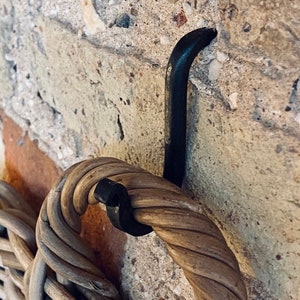 May include: Close-up of a woven wicker basket handle secured by a black metal hook against a textured brick and stone wall. The basket handle is made of tightly wound, light brown wicker.