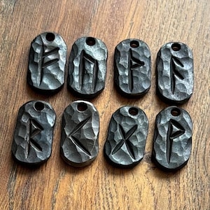 Può includere: Otto ciondoli in metallo nero con diverse rune incise. I ciondoli sono di forma ovale e hanno una texture martellata.