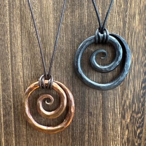 May include: Two pendants on dark cords, one crafted from hammered copper and the other from hammered dark iron. Each pendant features a circular outer ring with a spiral design extending toward the center, showcasing a textured, metallic finish.