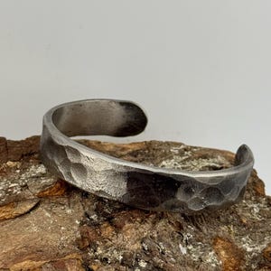 May include: A silver cuff bracelet with a hammered texture. The open-ended bracelet is curved and rests on a piece of wood with a rough, brown surface. The metal has a slightly darkened, aged appearance.