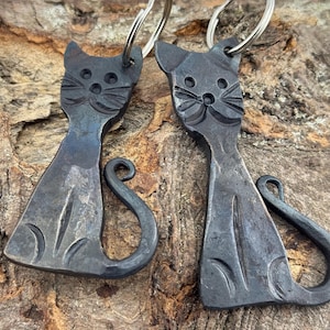 May include: Two handcrafted metal cat keychains. Each keychain features a stylized cat silhouette with etched details, including eyes, whiskers, and a curled tail. The keychains are attached to silver-colored key rings. The metal has a dark, textured finish.