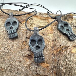 May include: Three dark grey, hand-forged metal skull pendants, each suspended from a thin black cord. The skull charms feature a textured, hammered finish with deep-set eye sockets, a triangular nasal cavity, and vertical lines representing teeth.