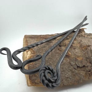 Könnte beinhalten: Drei handgeschmiedete Haarstäbe aus Metall mit einzigartigen Designs. Einer hat eine Spiralrose, ein anderer einen gedrehten Schaft und der dritte einen gebogenen Haken. Die dunkelgrauen Stäbe werden auf einem Holzstück präsentiert.