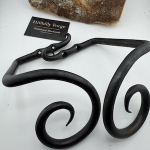 May include: Hand-forged, black metal decorative piece with a scroll design. The piece has a rectangular top with three holes and two curved ends. The text "Hillbilly Forge Traditional Blacksmith" is on a card.