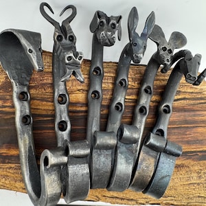 May include: Hand-forged metal wall hooks shaped like animals, including a horse, dog, rabbit, and goat. The hooks are mounted on a wooden base. The metal has a dark, textured finish. The sign at the bottom reads "Hillbilly Forge".