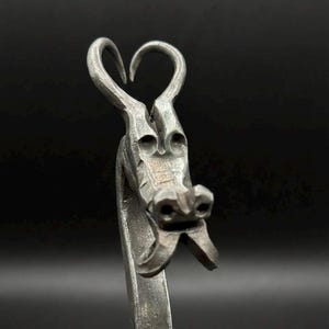 Könnte beinhalten: Handgefertigte Metallskulptur eines Drachenkopfes. Die Hörner des Drachen bilden eine Herzform. Die Skulptur besteht aus einem dunklen, metallischen Material mit sichtbaren Texturen und Details.