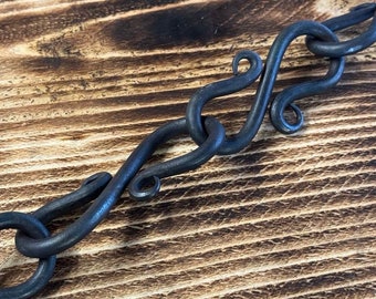 Puede incluir: Primer plano de una cadena de metal negro con eslabones decorativos forjados a mano. La cadena está sobre una superficie de madera.