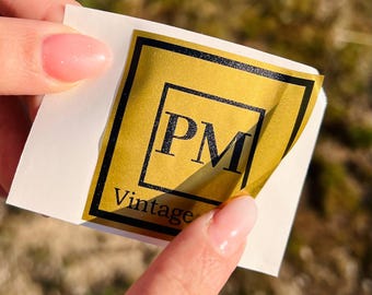 May include: A gold sticker with black text that reads "PM Vintage." The sticker has a black border and is being peeled from a white backing. The background is blurred.