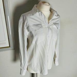 May include: A white button-down shirt with long sleeves and two chest pockets. The shirt is displayed on a mannequin. The shirt has a classic collar and button closures down the front. The fabric appears to be a lightweight material.