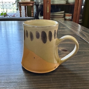 May include: A ceramic mug featuring a two-tone glaze with a light tan upper section decorated with a row of dark brown vertical oval spots and a solid peach-colored lower section. The mug has a curved handle and a smooth, rounded rim.
