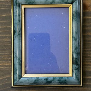 May include: A rectangular picture frame featuring a dark green faux marble finish with black edges and a thin gold inner border. The frame contains a solid purple insert with small white speckles scattered across the surface, creating a starry effect.
