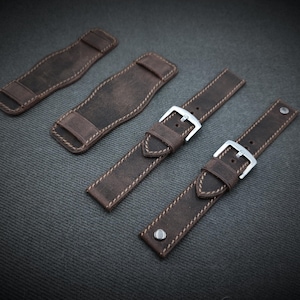 Può includere: Quattro cinturini e cuscinetti per orologi in pelle marrone sono disposti su una superficie grigio scuro. I cinturini hanno fibbie argentate e sono cuciti con filo di colore chiaro. I cuscinetti sono rettangolari con un passante.