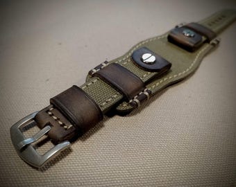 May include: A watch strap featuring a combination of olive green canvas and dark brown leather accents. The strap includes a silver-toned metal buckle, visible stitching along the edges, and two dark brown leather keepers with silver metal studs.
