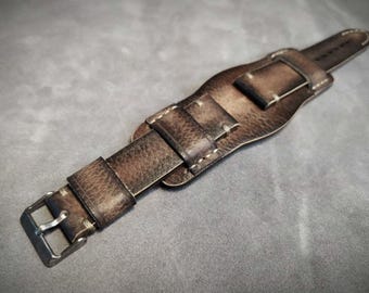 May include: A wide brown leather watch strap featuring a distressed finish with visible grain texture, light-coloured stitching, and a silver-toned metal buckle. The strap includes a central pad section designed to sit beneath a watch case.