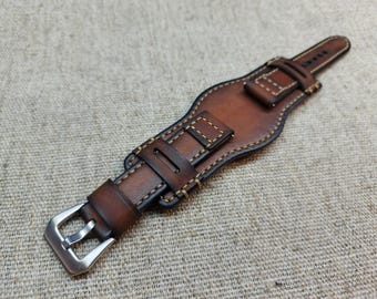 May include: A brown leather watch strap featuring a wide cuff design with visible tan stitching along the edges. The strap includes a silver-toned metal buckle and two leather keepers, showcasing a smooth, burnished finish on the surface.