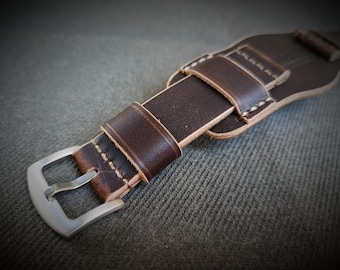 May include: Close-up of a brown leather watch strap with a silver buckle. The strap features white stitching and a padded design. The leather has a distressed look, suggesting a vintage or rugged style. The watch band is designed to fit a large watch.