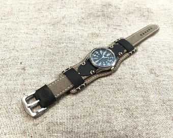 May include: A wristwatch featuring a round black analogue face with white numerals, set on a wide cuff-style strap. The strap combines tan canvas with black leather accents, secured by a silver-toned metal buckle and white contrast stitching.
