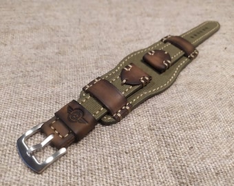 May include: An olive green canvas watch strap featuring brown leather accents and a silver-toned metal buckle. The strap includes decorative stitching and a stamped emblem on the leather loop, showcasing a winged design with a central circular motif.