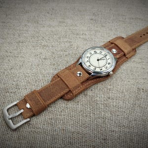 May include: A vintage-style wristwatch with a light-colored face and black numerals. The watch is set in a silver-toned case and is attached to a wide, brown leather cuff-style band. The band has a buckle closure and is secured with rivets.