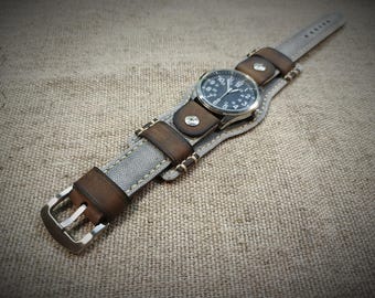 May include: A wristwatch featuring a round black analog face with white numerals, set into a wide cuff-style strap. The strap combines light gray canvas fabric with dark brown leather accents, silver-tone metal rivets, and a silver-tone buckle.