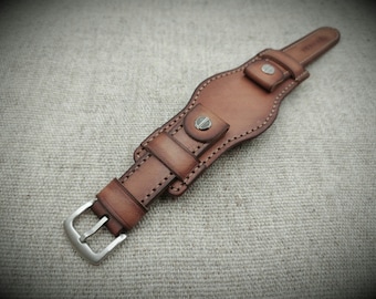 May include: A brown leather watch strap featuring a wide cuff design with visible stitching, a silver-toned metal buckle, and two metallic studs securing the central leather overlay. The leather has a gradient finish with darker edges.