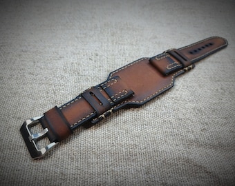May include: A brown leather watch strap featuring a wide cuff design with a gradient finish, visible light-colored stitching, and a silver-toned metal buckle. The leather displays a smooth texture with darker edges and a central rectangular panel.