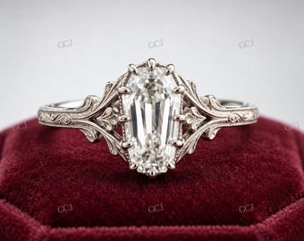 May include: A silver-coloured ring featuring a central coffin-shaped clear gemstone held by multiple prongs. The band and setting are decorated with intricate, raised scrollwork and filigree patterns that extend from the centre stone along the shank.