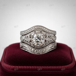 May include: A three-piece engagement ring set with a large, square-cut diamond center stone. The rings are silver-toned with intricate filigree and small accent diamonds. The set rests on a burgundy velvet ring box.