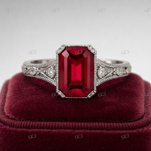 以下が含まれることがあります： 長方形の深い赤色の宝石が付いたシルバーリング。リングには、バンドと中央の石の周りに、詳細なフィリグリーと小さなダイヤモンドがあります。リングは赤いベルベットの表面に表示されています。