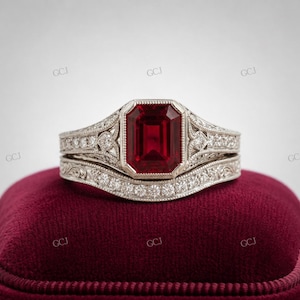 May include: A silver-toned ring set featuring an emerald-cut deep red gemstone as the centerpiece. The band and the setting are decorated with intricate filigree metalwork and small, clear pavé-set stones along the shank and the halo.