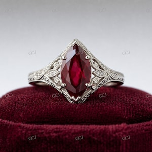 Könnte beinhalten: Ein Verlobungsring mit einem Rubin im Marquiseschliff und einem detaillierten, verzierten Silberband. Der Rubin hat eine tiefrote Farbe und ist in einer Fassung im Vintage-Stil mit zarten Filigranarbeiten und kleinen Akzentsteinen gefasst. Der Ring wird auf rotem Samt präsentiert.