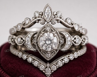 May include: A three-piece silver ring set with intricate detailing. The engagement ring features a large, round, clear stone surrounded by smaller accent stones. The bands have a vintage, ornate design with additional small stones, set on a burgundy velvet surface.