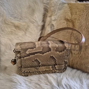 Könnte beinhalten: Eine rechteckige Umhängetasche mit strukturiertem Schlangenledermuster in Beige-, Hellbraun- und Dunkelbrauntönen. Die Tasche verfügt über einen strukturierten Klappverschluss, einen dünnen hellbraunen Lederriemen und kleine Metallbeschläge.