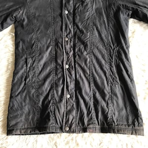 May include: A black jacket featuring a central vertical placket with four visible metal snap buttons. The garment includes two side pockets and vertical seam detailing running down the front panels, with a straight hemline at the bottom.