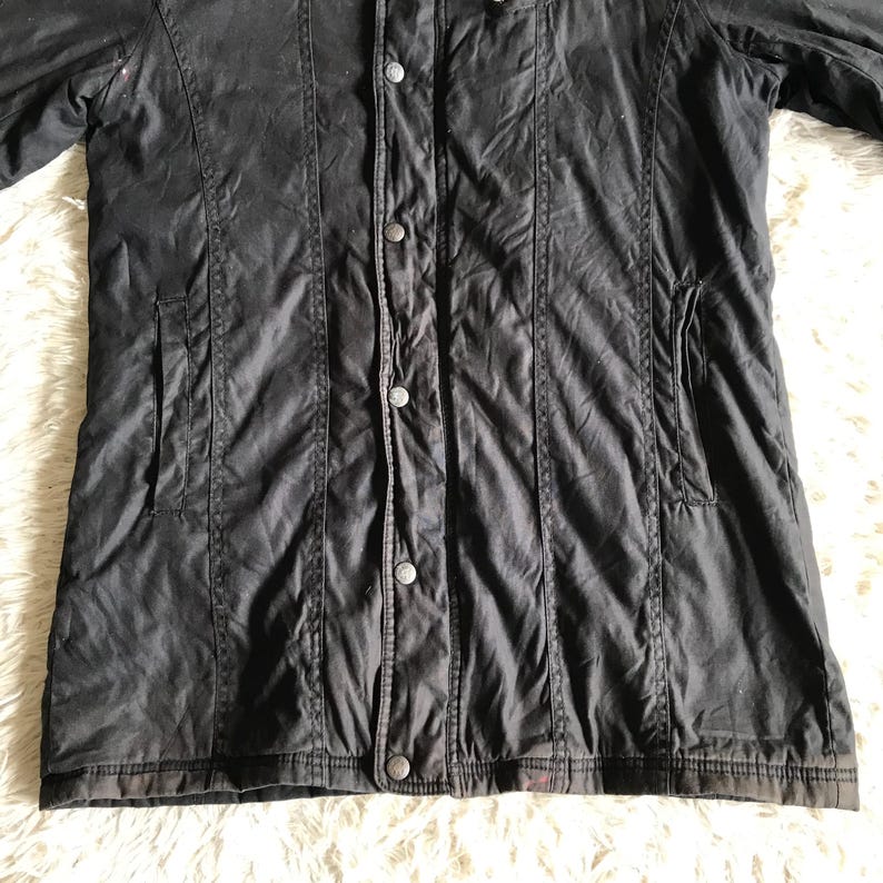 May include: A black jacket featuring a central vertical placket with four visible metal snap buttons. The garment includes two side pockets and vertical seam detailing running down the front panels, with a straight hemline at the bottom.
