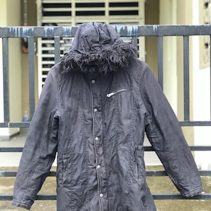 May include: A dark charcoal gray hooded jacket featuring a faux fur trim along the hood edge, a front button placket with visible silver-toned snap closures, long sleeves, and a single horizontal zippered pocket on the upper left chest area.