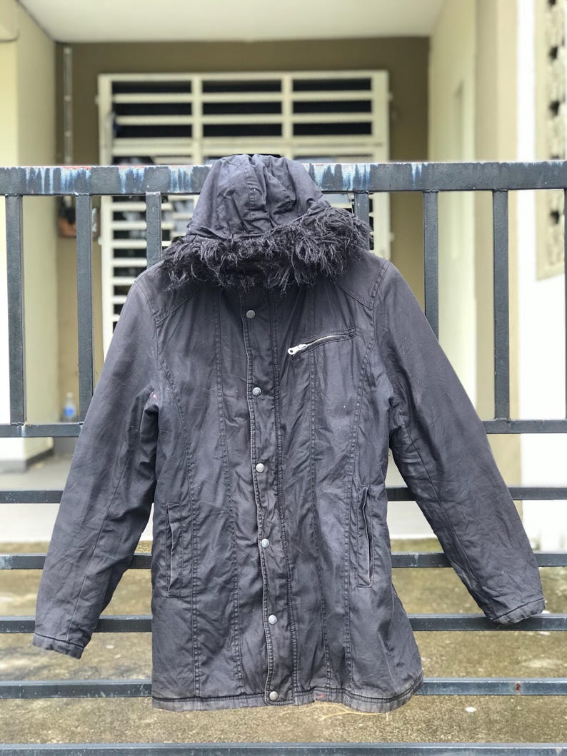 May include: A dark charcoal gray hooded jacket featuring a faux fur trim along the hood edge, a front button placket with visible silver-toned snap closures, long sleeves, and a single horizontal zippered pocket on the upper left chest area.