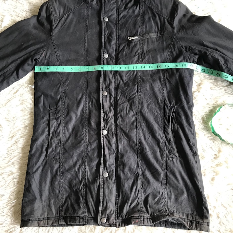 May include: A black jacket with a front button closure, a zippered chest pocket, and two side pockets. A green measuring tape is placed across the chest, showing a width of approximately 20 inches from side to side across the front panel.