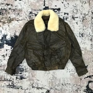 Könnte beinhalten: Braune Leder-Bomberjacke mit cremefarbenem Shearling-Kragen. Die Jacke hat zwei Vordertaschen mit Knopfverschlüssen und einen gerippten Saum. Das Leder hat ein Used-Aussehen.